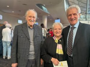 Three smiling seniors pose together in a bright event lobby: a man in a gray blazer, a woman in a black coat with scarf, and a man in a dark suit with a striped tie.