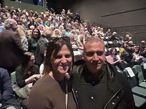Smiling couple posing for a photo in a crowded theater with hundreds of audience members in the background.