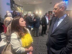 Woman in a yellow blouse talks with a suited man in a crowded lobby as other attendees mingle in the background.