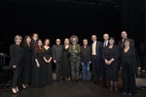Group portrait of about fourteen adults in black formal attire standing in a row on a dimly lit stage with a dark curtain backdrop, posing for a photo together.