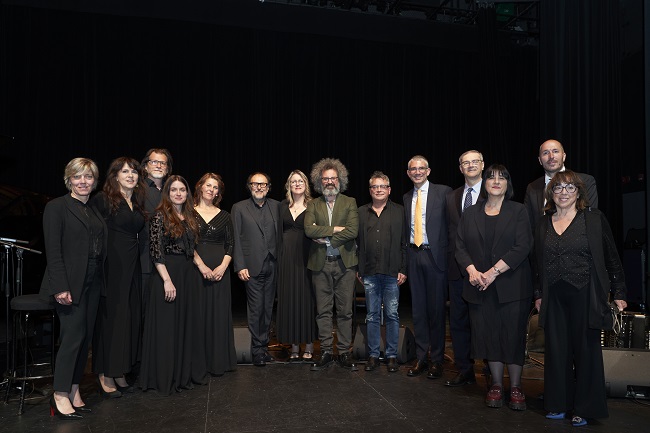 Group portrait of about fourteen adults in black formal attire standing in a row on a dimly lit stage with a dark curtain backdrop, posing for a photo together.