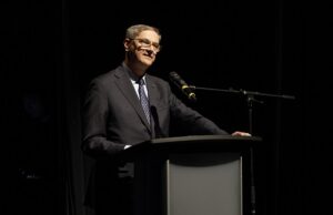 Man in a dark suit delivering a speech at a podium on a stage with a microphone there.