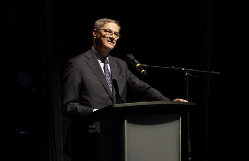 Man in a dark suit delivering a speech at a podium on a stage with a microphone there.