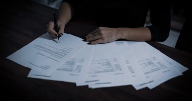 Person signs several documents at a wooden desk, pen in hand, papers spread out in front of them.