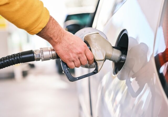Person refueling a car at a gas station, gripping a fuel nozzle near the fuel tank cap on the car's side