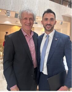 Two men in suits smiling for a photo in a modern lobby; older man with gray hair in a dark blazer and red plaid shirt, younger man in a blue suit holding a folder.