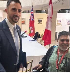 Two men at an indoor event near a long table with chairs, one standing in a suit holding a folder and the other seated with a red lanyard, flags in the background.
