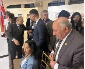 Group of formally dressed people networking at a reception; a man in a gray suit shakes hands with another man while a woman in blue sits nearby, flag visible in the background.