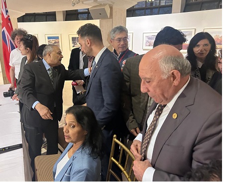 Group of formally dressed people networking at a reception; a man in a gray suit shakes hands with another man while a woman in blue sits nearby, flag visible in the background.
