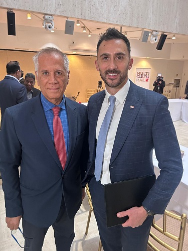 Two men in suits posing for a photo at a formal event; the older man wears a navy suit with a red tie, the younger man in a blue checked suit holds a black folder.