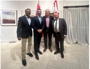 Four men in suits posing for a group photo at an indoor event, two shaking hands, with Canadian flags and artwork visible in the background.
