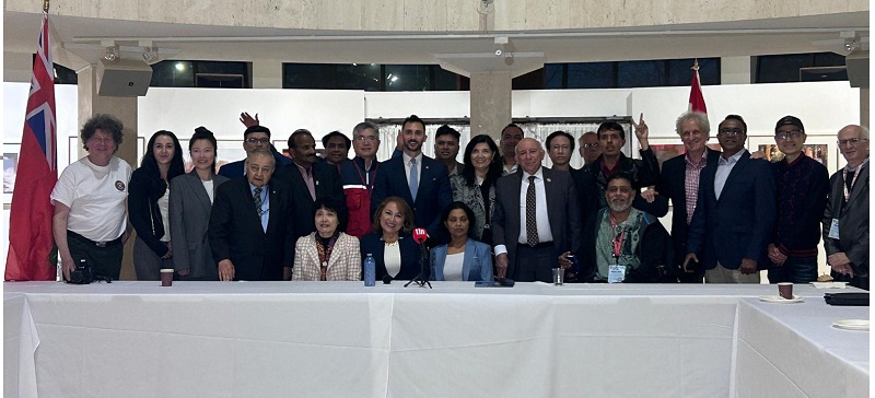 Diverse group of people standing behind a long white conference table in an indoor venue, with flags visible, posing for a group photo.