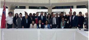 Group of diverse officials and attendees posing for a conference photo behind a long white table with microphones, flags on either side.