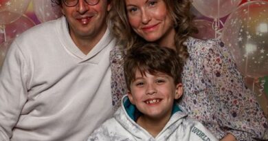 Family of three smiling at the camera in front of colorful balloons (father with glasses, mother in a floral blouse, son in a hoodie).