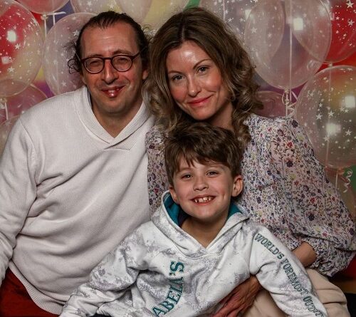 Family of three smiling at the camera in front of colorful balloons (father with glasses, mother in a floral blouse, son in a hoodie).
