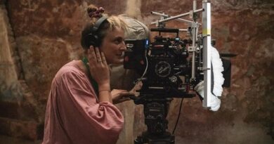 Woman in a pink blouse wearing headphones, operating a professional film camera on set on a tripod