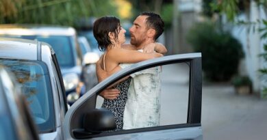 Couple embracing beside an open car door on a sunlit street, smiling at each other as they say goodbye or hello.