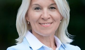 Professional woman smiling outdoors in a light blue blazer and white shirt, head-and-shoulders portrait.