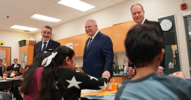 Three men in suits stand at the front of a classroom as students work at tables nearby during a school visit.