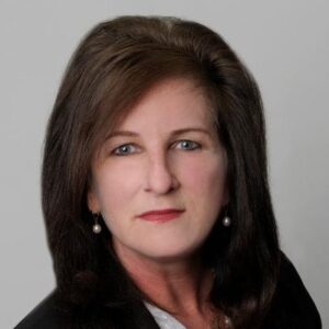 Professional headshot of a middle-aged woman with dark brown hair, pearl earrings, and a black blazer.