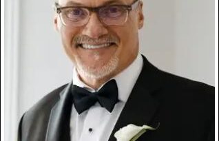 Smiling man in a black tuxedo with bow tie and boutonniere, posing for a formal portrait.