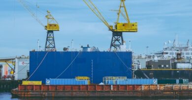 Large blue cargo module on a barge with two yellow gantry cranes at a shipyard by the water, under a clear sky.