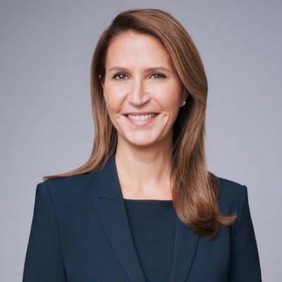 Professional headshot of a smiling woman in a navy blazer against a gray studio backdrop.