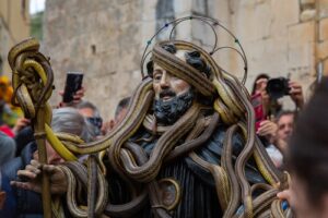 Performer in a Medusa-inspired costume with many snake props and a circular halo, crowd and photographers in background.