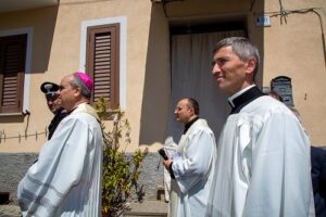 Three priests in white liturgical robes walk in a procession along a sunlit street outside a beige building; the bishop wears a pink zucchetto.