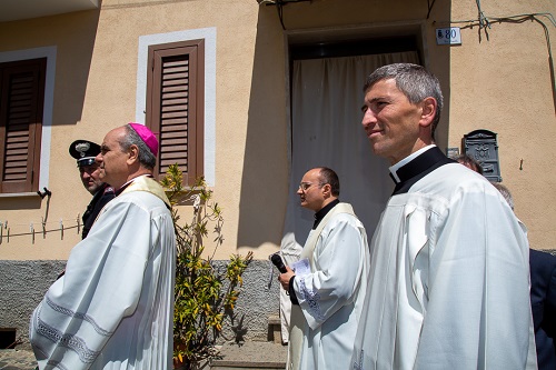 Three priests in white liturgical robes walk in a procession along a sunlit street outside a beige building; the bishop wears a pink zucchetto.