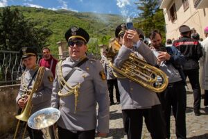 Police band members in gray uniforms play brass instruments outdoors, with mountains visible in the background and people photographing the scene.