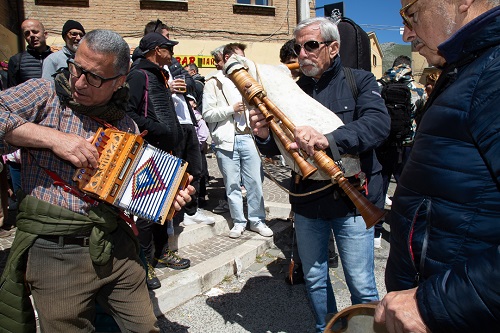 Outdoor street gathering: a man in a plaid shirt plays an accordion while another man with sunglasses plays a wooden bagpipe, surrounded by onlookers.