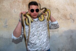 Man in sunglasses and a patterned shirt with multiple snakes draped over his shoulders against a beige wall.