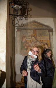 Woman with blonde hair wearing a white face mask and scarf stands indoors in front of a faded religious mural; another woman in a dark jacket is nearby.