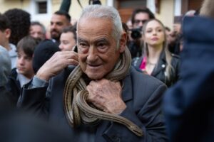 Older man in a navy jacket adjusts a brown scarf while surrounded by a crowd at a public event.