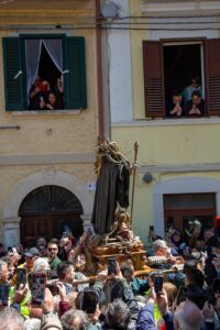 Religious statue being carried on a platform through a crowded street as spectators film with smartphones; colorful buildings and people watching from windows.