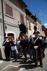 Religious procession on a cobblestone street; a gilded statue is carried by men, flanked by uniformed guards with plumed hats.