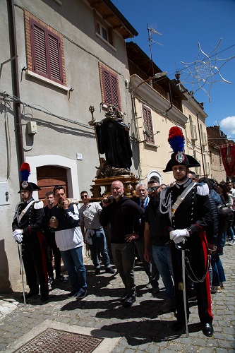 Religious procession on a cobblestone street; a gilded statue is carried by men, flanked by uniformed guards with plumed hats.