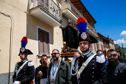 Men in ceremonial uniforms with plumed hats carry a statue on a wooden platform during a sunny street procession.