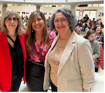 Three women posing together at an indoor event, smiling, with a crowd in the background.