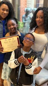 Smiling woman and three children pose indoors; a girl in front holds a certificate.