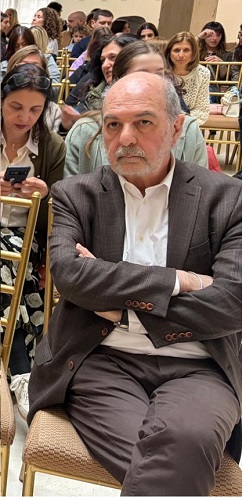 Older man in a brown blazer sits in the front row with arms crossed at a formal event, surrounded by seated attendees.