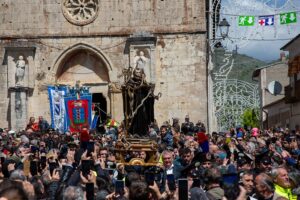 Statue of a saint carried on a platform through a crowded stone church square during a festival, with banners and onlookers surrounding it.