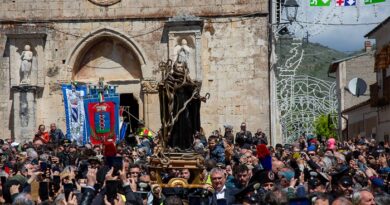 Statue of a saint carried on a platform through a crowded stone church square during a festival, with banners and onlookers surrounding it.