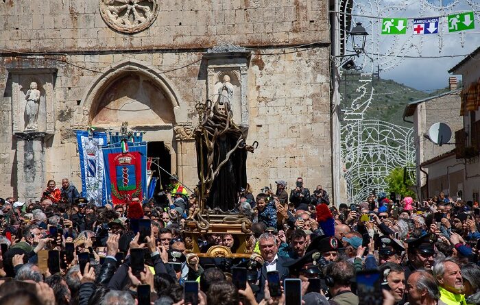 Statue of a saint carried on a platform through a crowded stone church square during a festival, with banners and onlookers surrounding it.