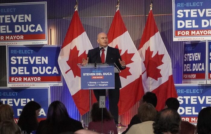 Politician Steven Del Duca giving a campaign speech at a podium with Canadian flags and multiple 'Re-Elect Steven Del Duca for Mayor' signs behind him.