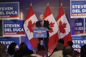 A man giving a campaign speech at a podium, flanked by multiple Canadian flags and blue 'Re-Elect for Mayor' signs behind him on stage.