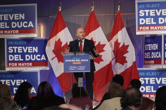 A man giving a campaign speech at a podium, flanked by multiple Canadian flags and blue 'Re-Elect for Mayor' signs behind him on stage.