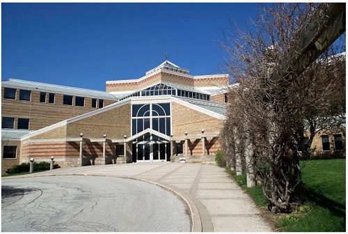 Front view of a large brick campus building with a glass entryway and curved driveway under a clear blue sky.