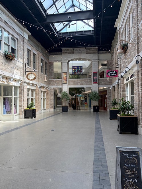 Indoor shopping corridor with brick storefronts, a glass skylight, string lights, and potted plants along a wide walkway on a mall concourse.